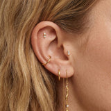 Detailed close-up of a blonde woman’s ear wearing multiple gold earrings, including a gold ear cuff, small gold hoops, a baguette-style stud, and a long gold threader earring with a sculptural wave and crystal accent. Ideal for women with multiple piercings, this image demonstrates how the gold threader earrings can be styled with hoops and cuffs for a curated, modern ear stack.