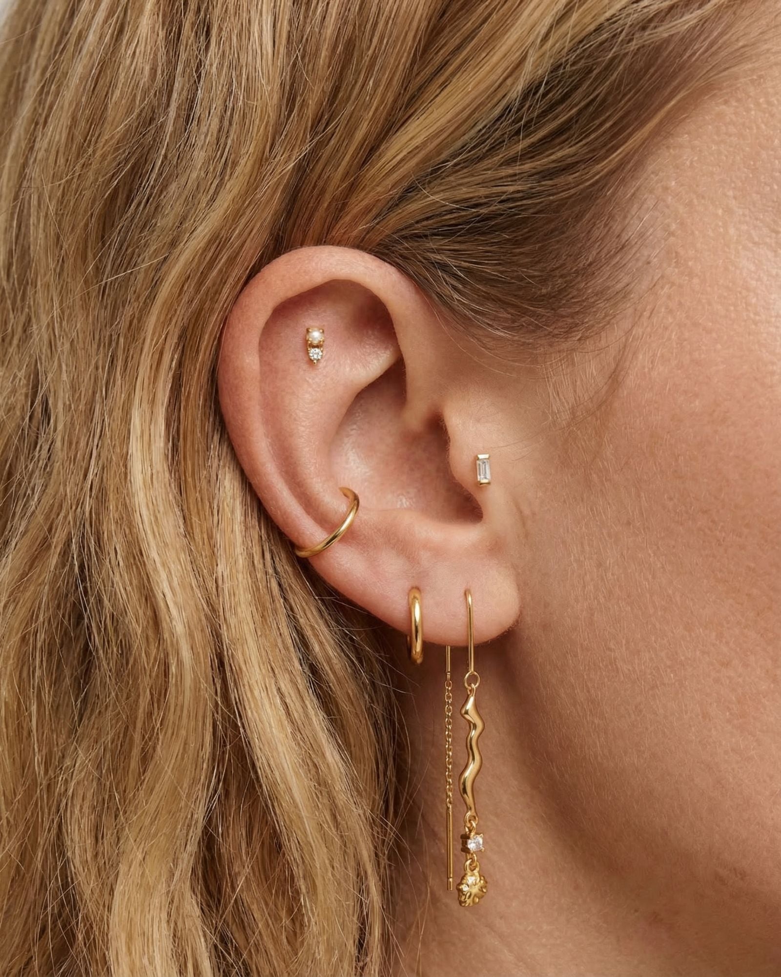 Detailed close-up of a blonde woman’s ear wearing multiple gold earrings, including a gold ear cuff, small gold hoops, a baguette-style stud, and a long gold threader earring with a sculptural wave and crystal accent. Ideal for women with multiple piercings, this image demonstrates how the gold threader earrings can be styled with hoops and cuffs for a curated, modern ear stack.