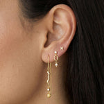 Close-up side profile of a woman’s ear wearing a layered gold earring stack, featuring delicate gold stud earrings in the upper ear, a small gold drop earring with a pearl accent, and a long gold drop earring with a sculptural wavy design and fine chain detail. The image clearly shows the varied lengths, textures, and movement of the earrings when worn together. Designed for women who enjoy refined yet expressive ear styling, this image helps customers visualise how to mix gold studs and drop earrings to cr