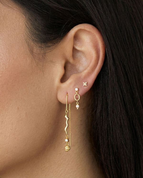 Close-up side profile of a woman’s ear wearing a layered gold earring stack, featuring delicate gold stud earrings in the upper ear, a small gold drop earring with a pearl accent, and a long gold drop earring with a sculptural wavy design and fine chain detail. The image clearly shows the varied lengths, textures, and movement of the earrings when worn together. Designed for women who enjoy refined yet expressive ear styling, this image helps customers visualise how to mix gold studs and drop earrings to cr