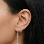 Side profile close-up of a dark-haired woman’s ear wearing silver earrings, featuring a pavé huggie hoop, a small cartilage stud, and a long silver chain threader earring with a wavy bar and crystal drop. Created for women who prefer refined silver jewellery, this image helps shoppers see proportion, sparkle, and how the silver drop earrings complement other minimalist ear piercings.