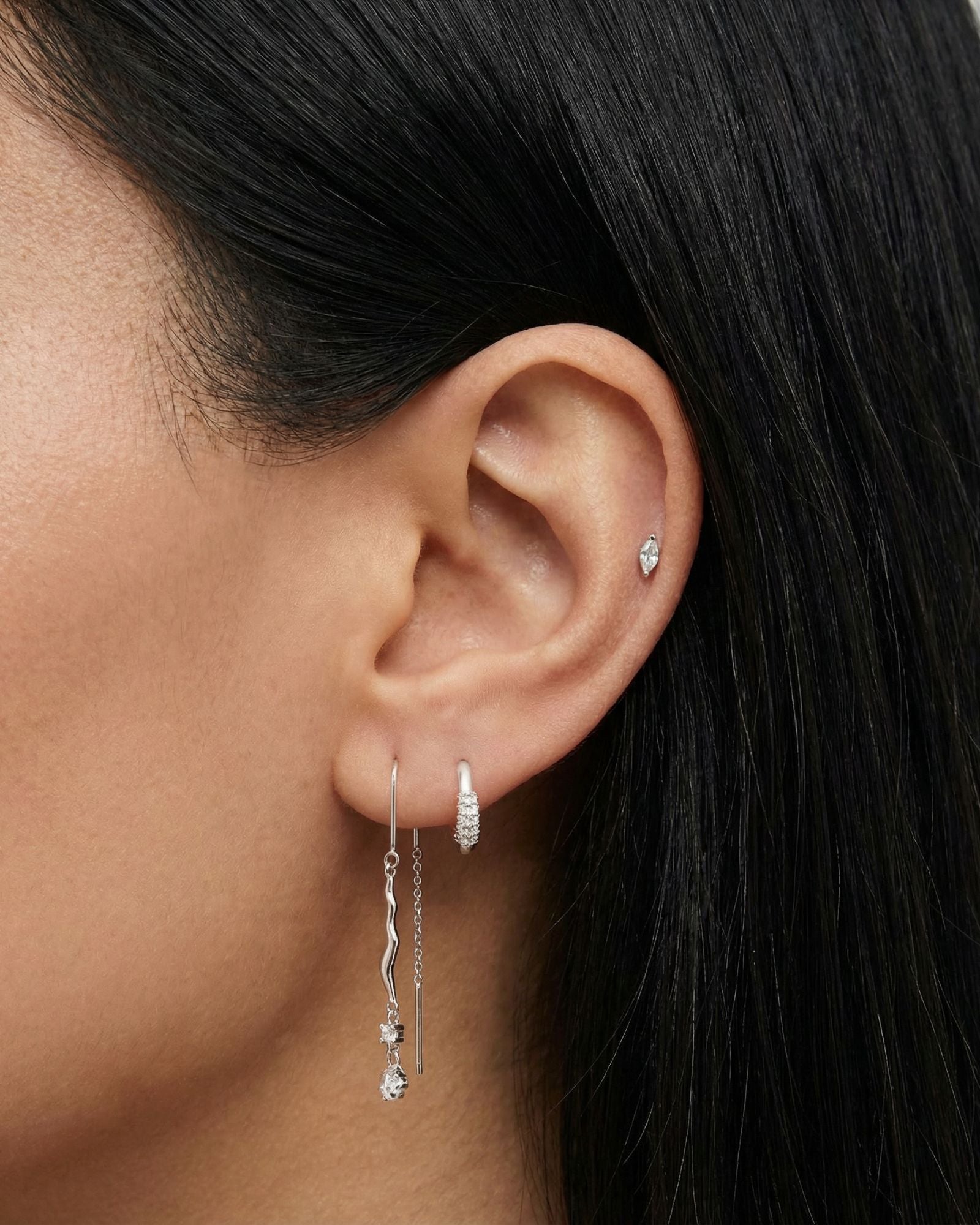 Side profile close-up of a dark-haired woman’s ear wearing silver earrings, featuring a pavé huggie hoop, a small cartilage stud, and a long silver chain threader earring with a wavy bar and crystal drop. Created for women who prefer refined silver jewellery, this image helps shoppers see proportion, sparkle, and how the silver drop earrings complement other minimalist ear piercings.