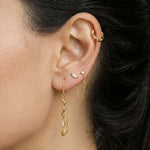 Close-up side profile of a woman’s ear wearing a curated gold ear stack, including a slim gold ear cuff on the cartilage, delicate gold stud earrings, a fine gold chain connector, and a long gold drop earring with a sculptural wavy detail and small stone accent. The image clearly shows the layered styling, varied lengths, and movement of the drop earring against natural skin and dark hair. Designed for women who enjoy expressive ear styling, this image helps customers visualise how to combine gold studs
