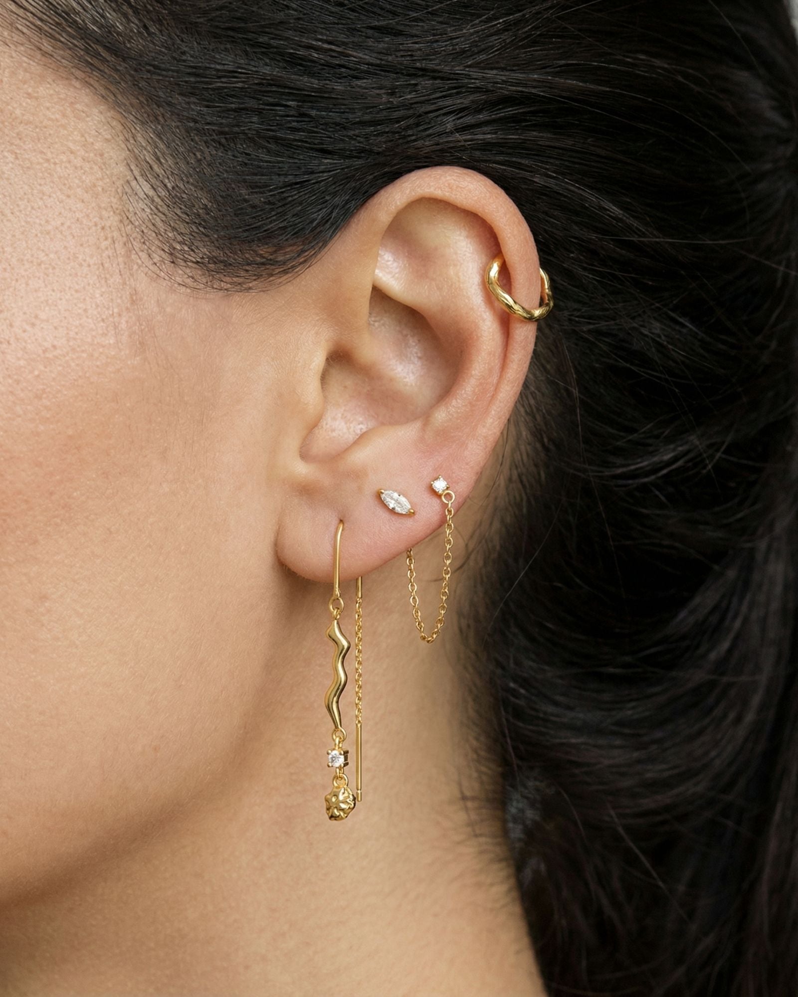 Close-up side profile of a woman’s ear wearing a curated gold ear stack, including a slim gold ear cuff on the cartilage, delicate gold stud earrings, a fine gold chain connector, and a long gold drop earring with a sculptural wavy detail and small stone accent. The image clearly shows the layered styling, varied lengths, and movement of the drop earring against natural skin and dark hair. Designed for women who enjoy expressive ear styling, this image helps customers visualise how to combine gold studs