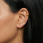 Close-up product photo of a woman’s ear with warm medium skin tone and sleek dark hair, styled with layered gold earrings across multiple piercings. The ear features a small textured gold hoop in the upper cartilage, a tiny gold crescent moon stud and a small star-shaped stud in the mid ear, and a delicate gold drop earring in the lobe with a wavy bar, fine chain, and small clear stone detail. This image is for customers looking for expressive gold earrings that combine minimal celestial motifs with elegant