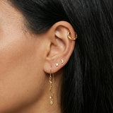 Close-up product photo of a woman’s ear with warm medium skin tone and sleek dark hair, styled with layered gold earrings across multiple piercings. The ear features a small textured gold hoop in the upper cartilage, a tiny gold crescent moon stud and a small star-shaped stud in the mid ear, and a delicate gold drop earring in the lobe with a wavy bar, fine chain, and small clear stone detail. This image is for customers looking for expressive gold earrings that combine minimal celestial motifs with elegant