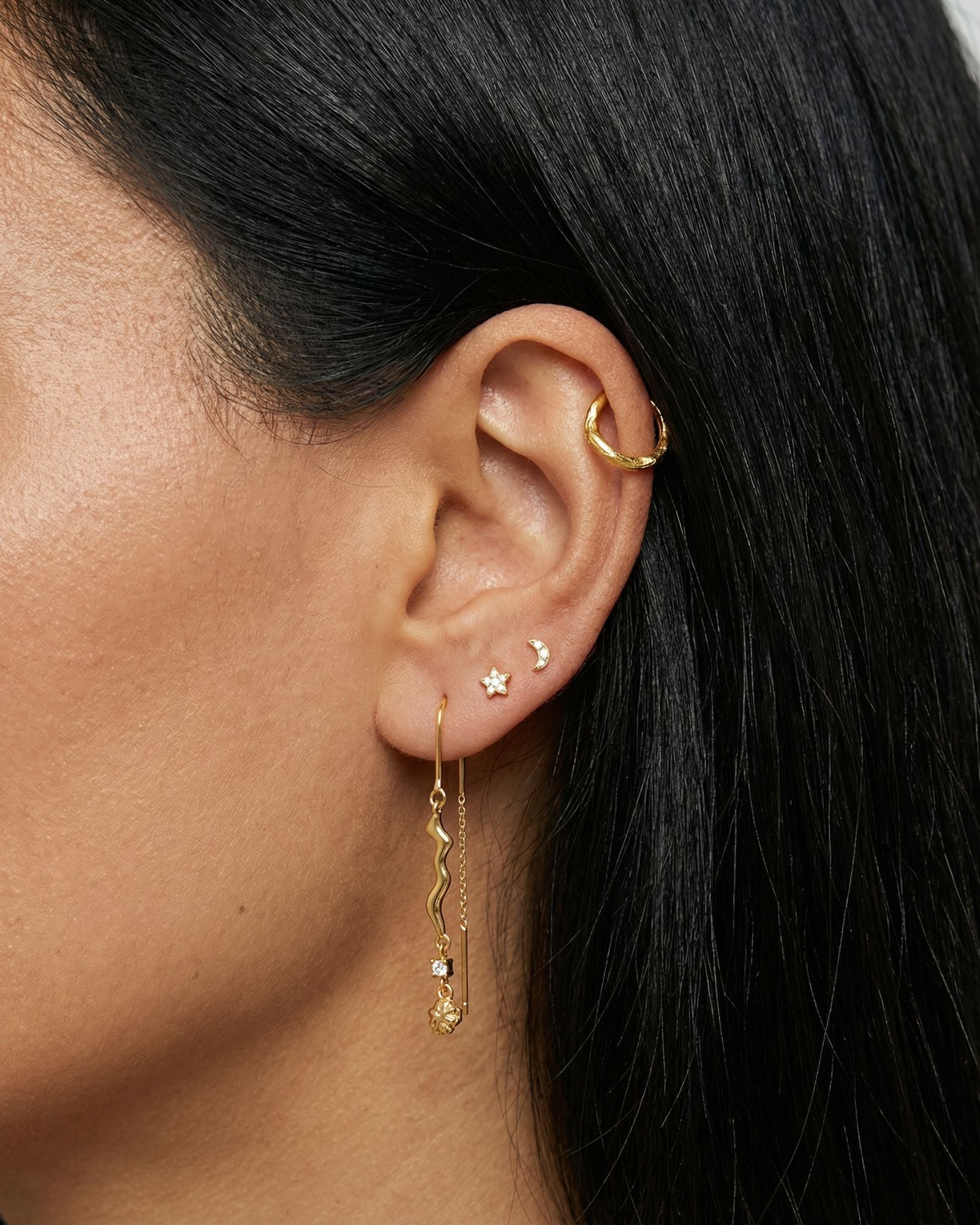 Close-up product photo of a woman’s ear with warm medium skin tone and sleek dark hair, styled with layered gold earrings across multiple piercings. The ear features a small textured gold hoop in the upper cartilage, a tiny gold crescent moon stud and a small star-shaped stud in the mid ear, and a delicate gold drop earring in the lobe with a wavy bar, fine chain, and small clear stone detail. This image is for customers looking for expressive gold earrings that combine minimal celestial motifs with elegant