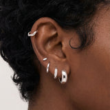 Close-up of a woman’s ear wearing silver Eva hoop earrings layered with small silver studs and cartilage jewellery. This image is for women searching for silver ear stack ideas and practical examples of how to style multiple piercings in a clean, modern way.

