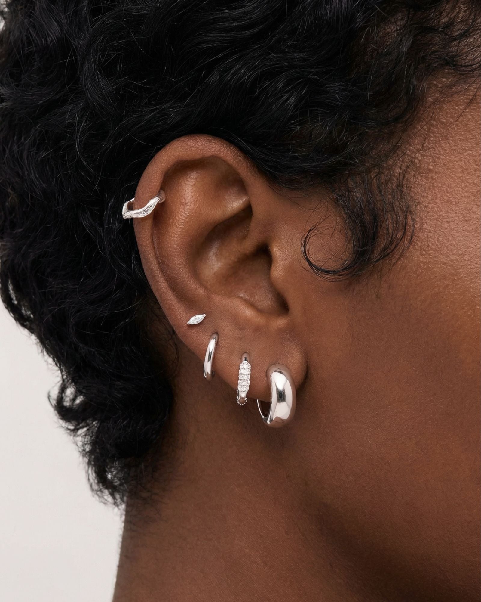 Close-up of a woman’s ear wearing silver Eva hoop earrings layered with small silver studs and cartilage jewellery. This image is for women searching for silver ear stack ideas and practical examples of how to style multiple piercings in a clean, modern way.
