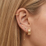 Close-up side profile of a blonde woman’s ear wearing a layered gold ear stack, including a smooth gold ear cuff, small gemstone studs, a slim gold chain drop earring with a bead detail, and a wide gold hoop. Designed for women with multiple piercings, this image helps customers see how gold stud earrings and drop chains can be styled together for a balanced, modern everyday ear stack.