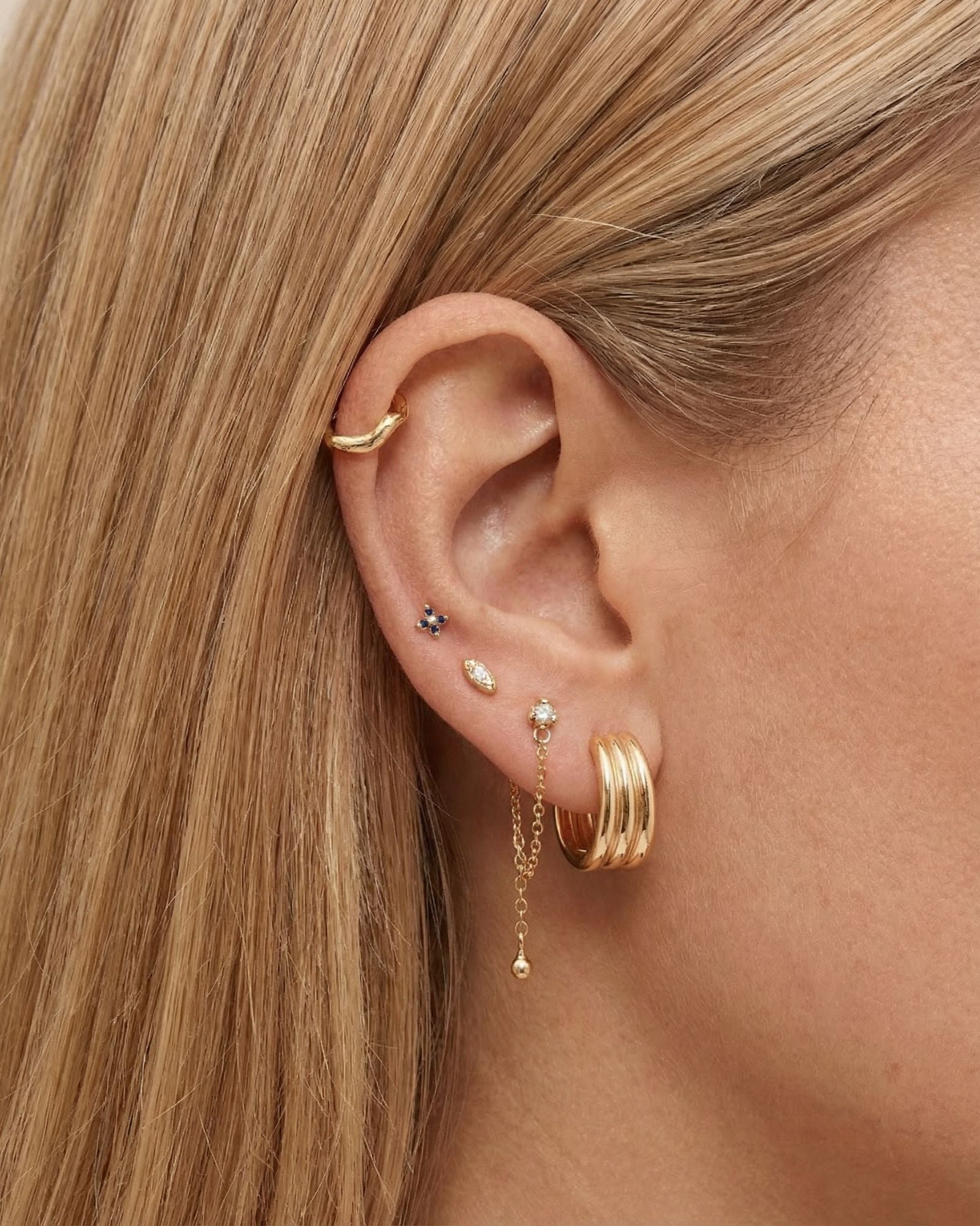 Close-up side profile of a blonde woman’s ear wearing a layered gold ear stack, including a smooth gold ear cuff, small gemstone studs, a slim gold chain drop earring with a bead detail, and a wide gold hoop. Designed for women with multiple piercings, this image helps customers see how gold stud earrings and drop chains can be styled together for a balanced, modern everyday ear stack.