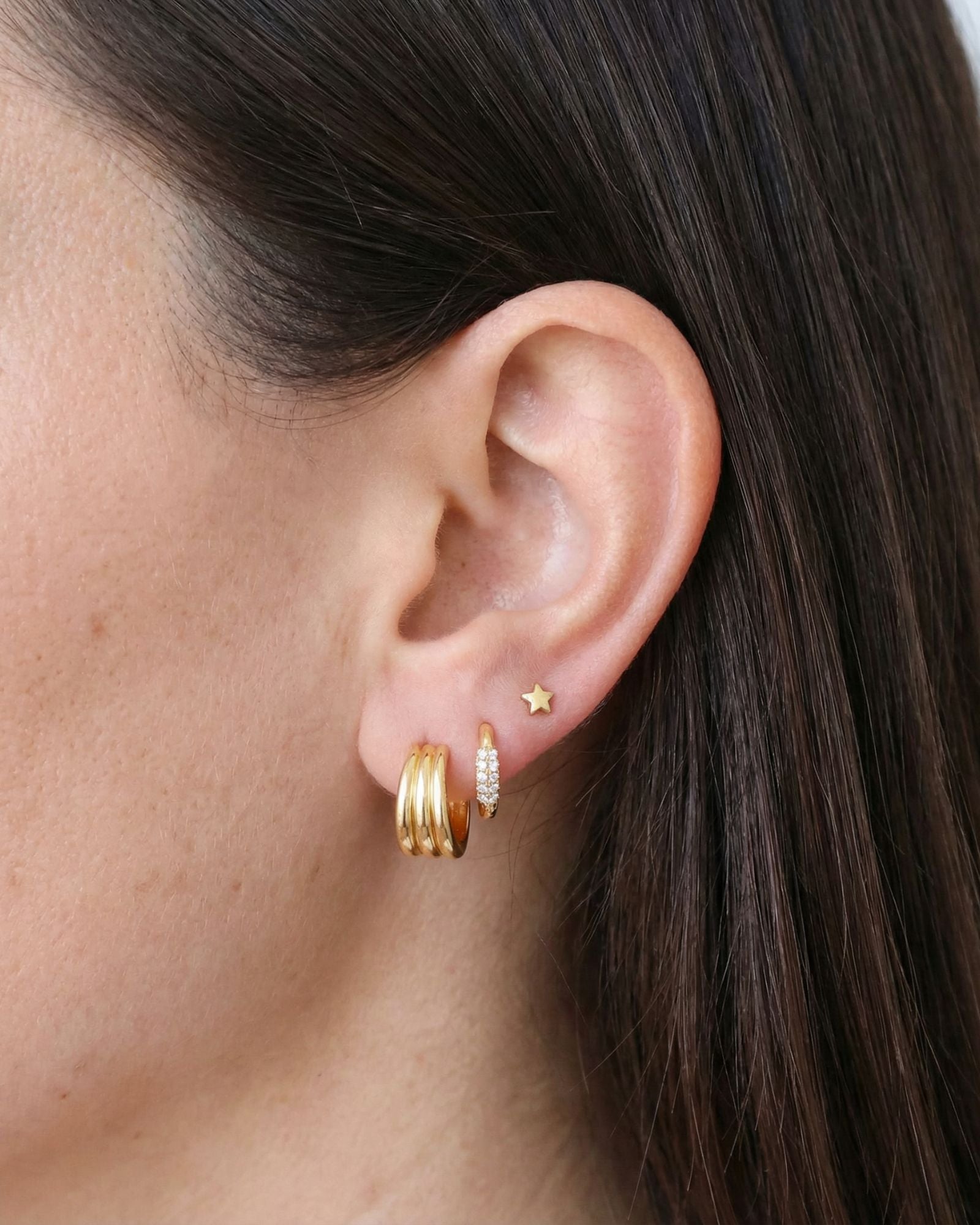 Close-up side profile of a brunette woman’s ear styled with gold jewellery, featuring stacked gold hoops, a pavé huggie earring, and a small gold star stud. Created for women who want refined gold earrings for daily wear, this image shows size, placement, and layering options to help customers choose gold hoops and huggies that work together without looking heavy or overcrowded.