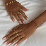 Product image of both hands resting on white fabric, wearing multiple gold rings including an organic spiral gold ring, slim stacking bands, and a signet-style ring, paired with gold bangles and a fine bracelet. Designed for women looking for everyday gold jewellery, this image shows how minimalist gold rings can be layered to create a cohesive, effortless, and versatile styling solution.