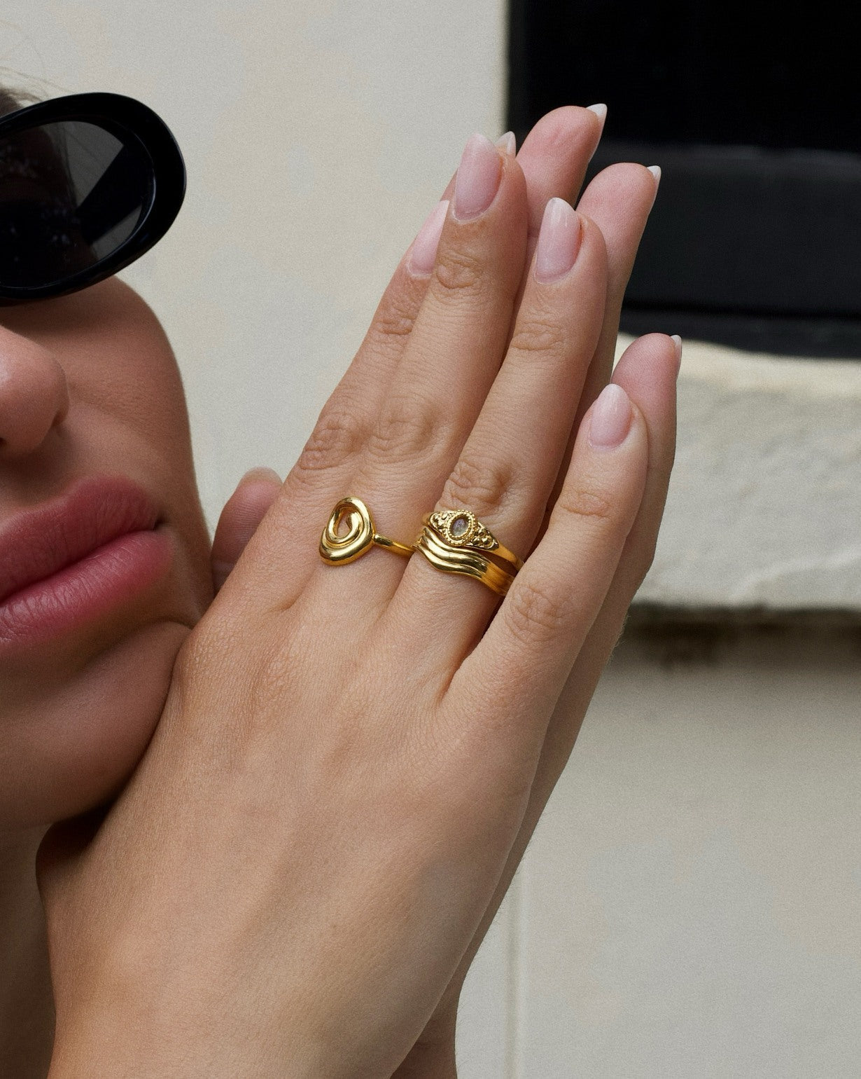 Product image showing a close-up of a hand wearing layered gold rings, including a sculptural heart-shaped gold ring and a wave-style gold stacking ring, styled on a model with neutral manicure and sunglasses. Designed for women looking for gold stacking rings, minimalist gold jewellery, and everyday statement rings, this image demonstrates how to layer gold rings for a cohesive, elevated look while solving the need for versatile pieces that transition easily from casual to refined wear.