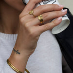 Product image showing a close-up of a hand wearing layered gold rings, double gold bangle, including a sculptural heart-shaped gold ring and a wave-style gold stacking ring, styled on a model with neutral manicure and sunglasses. Designed for women looking for gold stacking rings, minimalist gold jewellery, and everyday statement rings, this image demonstrates how to layer gold rings for a cohesive, elevated look while solving the need for versatile pieces that transition easily from casual to refined wear.