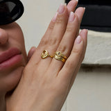Product image showing a close-up of a hand wearing layered gold rings, including a sculptural heart-shaped gold ring and a wave-style gold stacking ring, styled on a model with neutral manicure and sunglasses. Designed for women looking for gold stacking rings, minimalist gold jewellery, and everyday statement rings, this image demonstrates how to layer gold rings for a cohesive, elevated look while solving the need for versatile pieces that transition easily from casual to refined wear.
