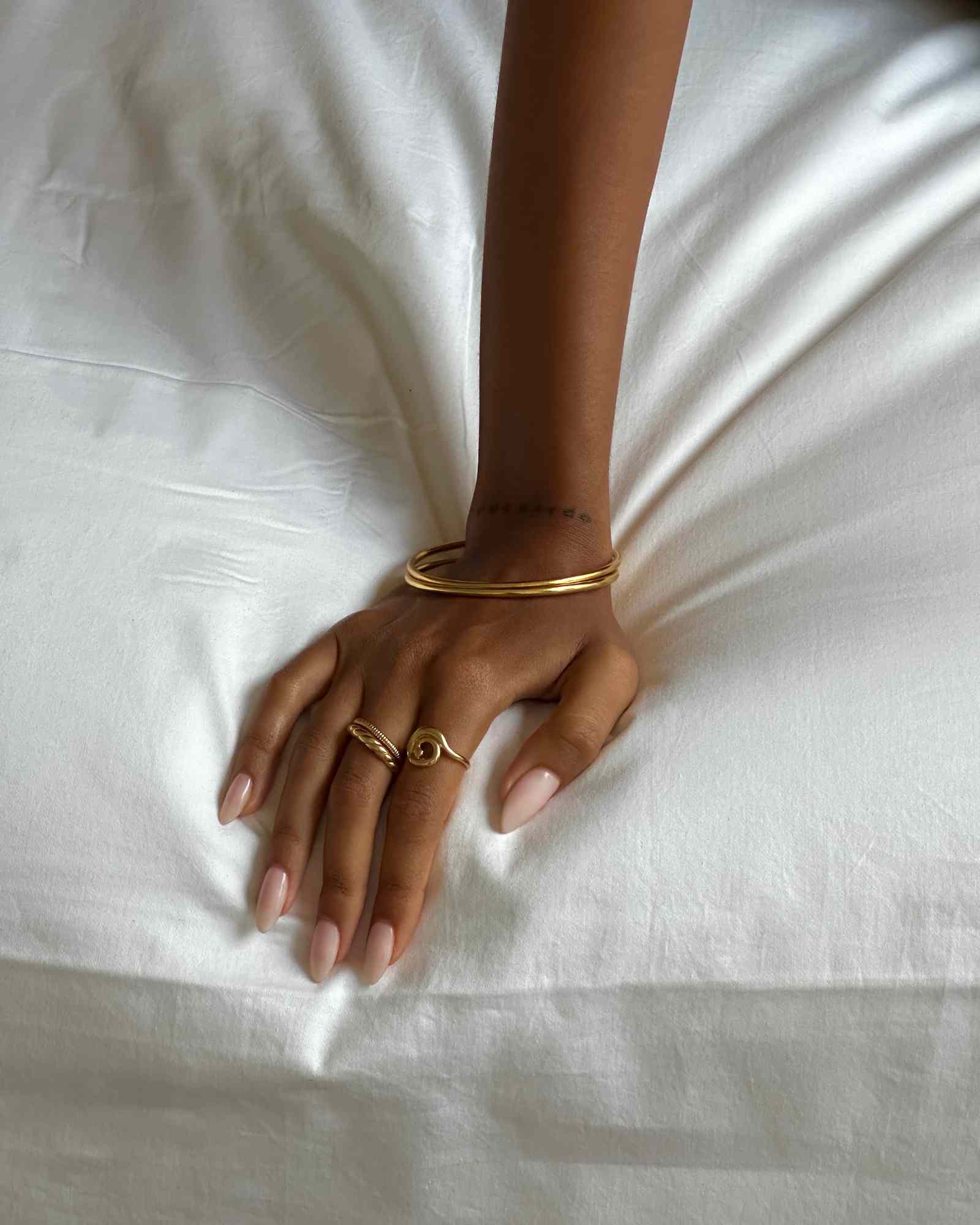 Product image of a woman’s hand resting on white fabric, wearing an organic spiral gold ring paired with a slim gold band and gold bangles. Designed for women seeking minimalist gold jewellery for everyday wear, this image highlights how a spiral gold stacking ring creates a refined, effortless look while remaining comfortable and easy to style daily.