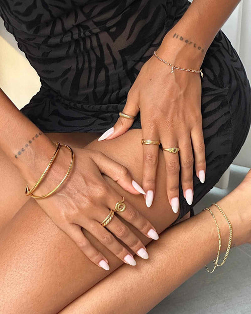 Product image of a woman seated wearing multiple gold rings including an organic spiral gold ring, a slim gold band, and a signet-style ring, paired with gold bangles and bracelets. Designed for women building a cohesive jewellery look, this image shows how everyday gold rings and minimalist gold jewellery can be stacked and styled for a polished yet effortless outfit.