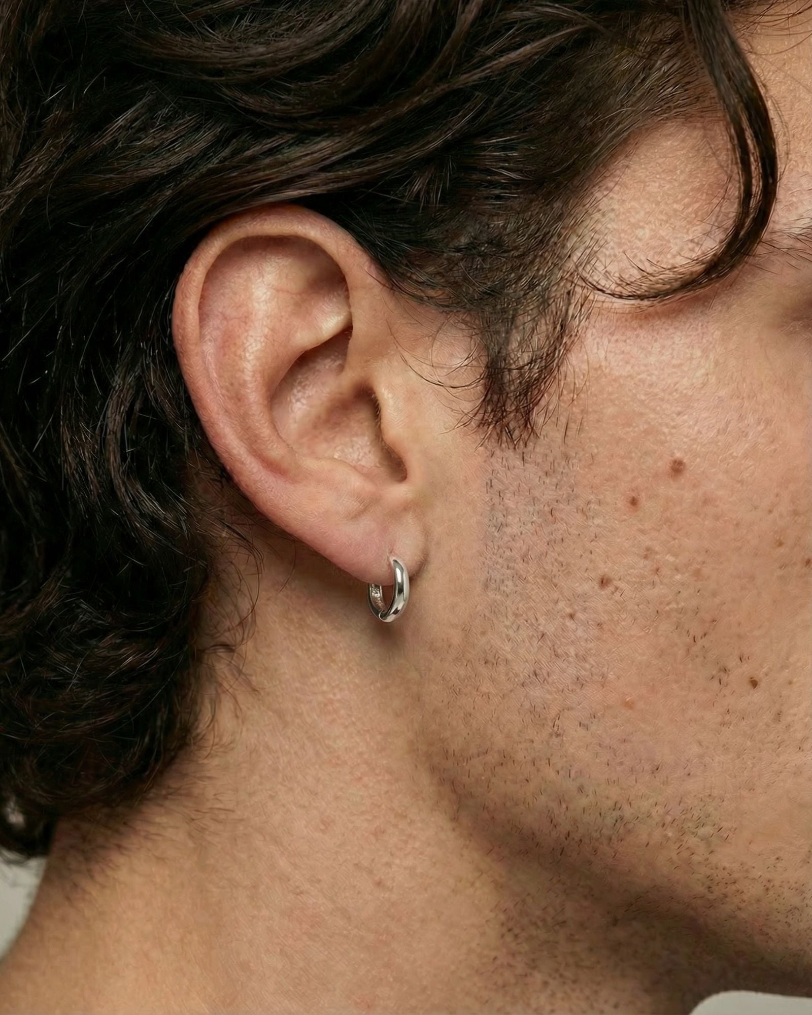Close-up side profile of a person wearing a small polished silver hoop earring in a pierced earlobe. The minimalist hoop sits snugly against the ear with a smooth, rounded finish, shown against natural skin texture and short facial hair. This image is for customers looking for a simple, everyday silver hoop earring that is lightweight, understated, and suitable for daily wear or layering with other jewellery.