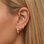 Close-up product photo of a woman’s ear with fair skin and straight blonde hair, styled with layered gold earrings. The ear features a smooth, medium-sized gold huggie hoop in the lobe, a small gold flower-shaped huggie set with clear stones in a second piercing, and a slim gold pavé huggie with clear stones in the upper lobe. This image is for customers looking for elegant gold earrings that add subtle sparkle while remaining suitable for everyday wear. It shows how delicate gold huggies with stones can be