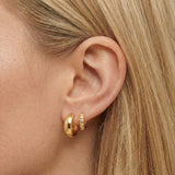 Close-up product photo of a woman’s ear with fair skin and straight blonde hair, styled with two layered gold huggie hoops in the lobe. The front piercing features a smooth, rounded gold huggie, while the second piercing features a smaller textured gold huggie with a subtle hammered finish. This image is for customers looking for simple gold earrings for everyday wear or a minimal ear stack. It shows how mixing smooth and textured gold huggies can add quiet detail and dimension while keeping a clean, unders