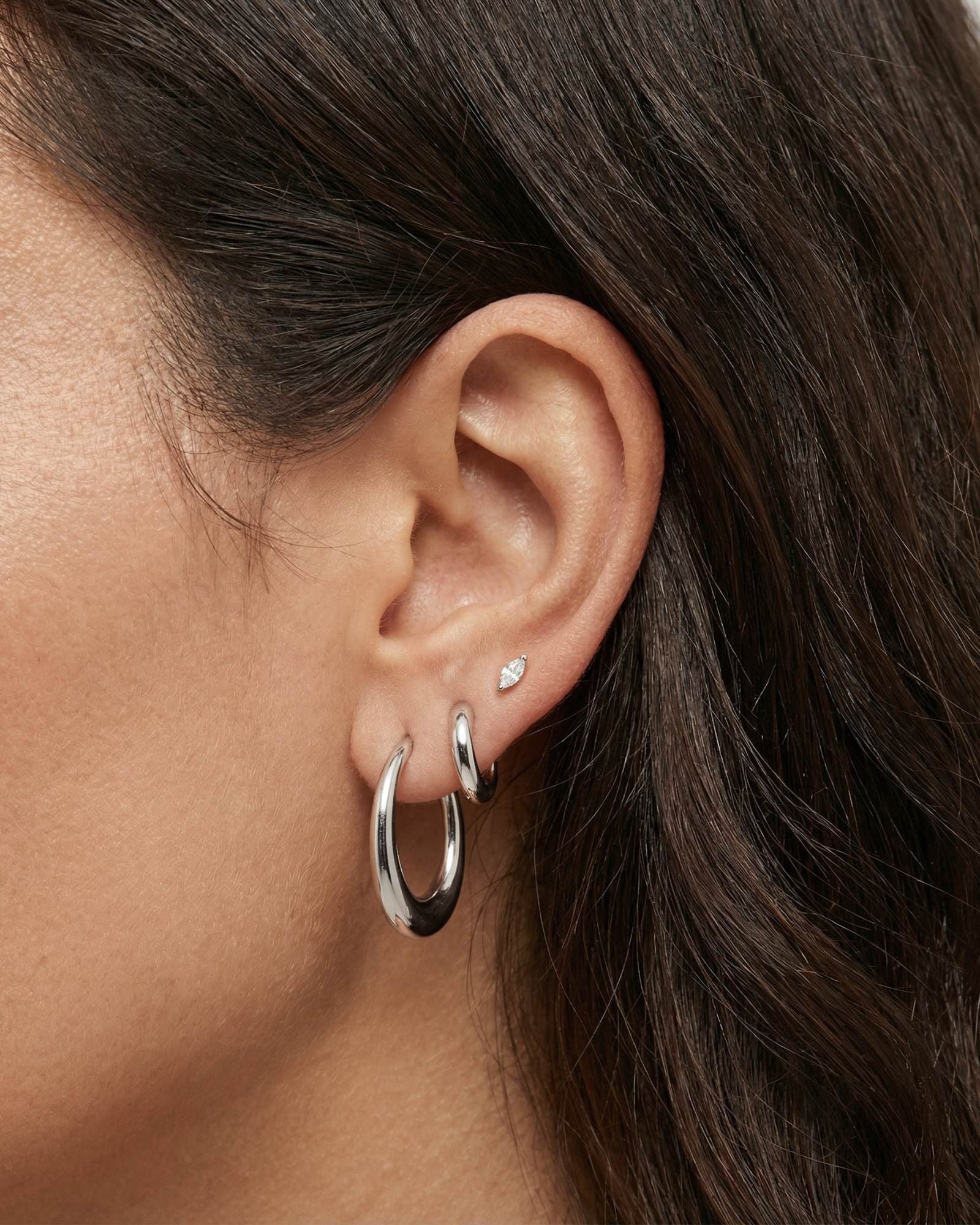 Close-up side profile of a woman’s ear wearing polished silver hoop earrings in two sizes, paired with a small silver marquise-shaped stud in the upper lobe. The image highlights the smooth, reflective finish and classic rounded shape of the silver hoops against natural skin and dark hair. Designed for women who want versatile, everyday silver earrings that are easy to style and suitable for multiple piercings, this image helps shoppers visualise scale, layering, and how minimal silver hoops look when worn.
