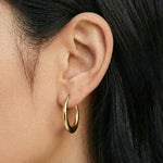 Close-up side profile of a woman’s ear wearing a single pair of polished gold hoop earrings with a rounded, medium-sized silhouette. The photo clearly shows the warm gold tone, clean finish, and proportion of the hoop on the ear against natural skin and dark hair. Designed for women seeking timeless, everyday gold hoop earrings, this image helps customers understand size, fit, and how classic gold hoops elevate a simple, minimal look.