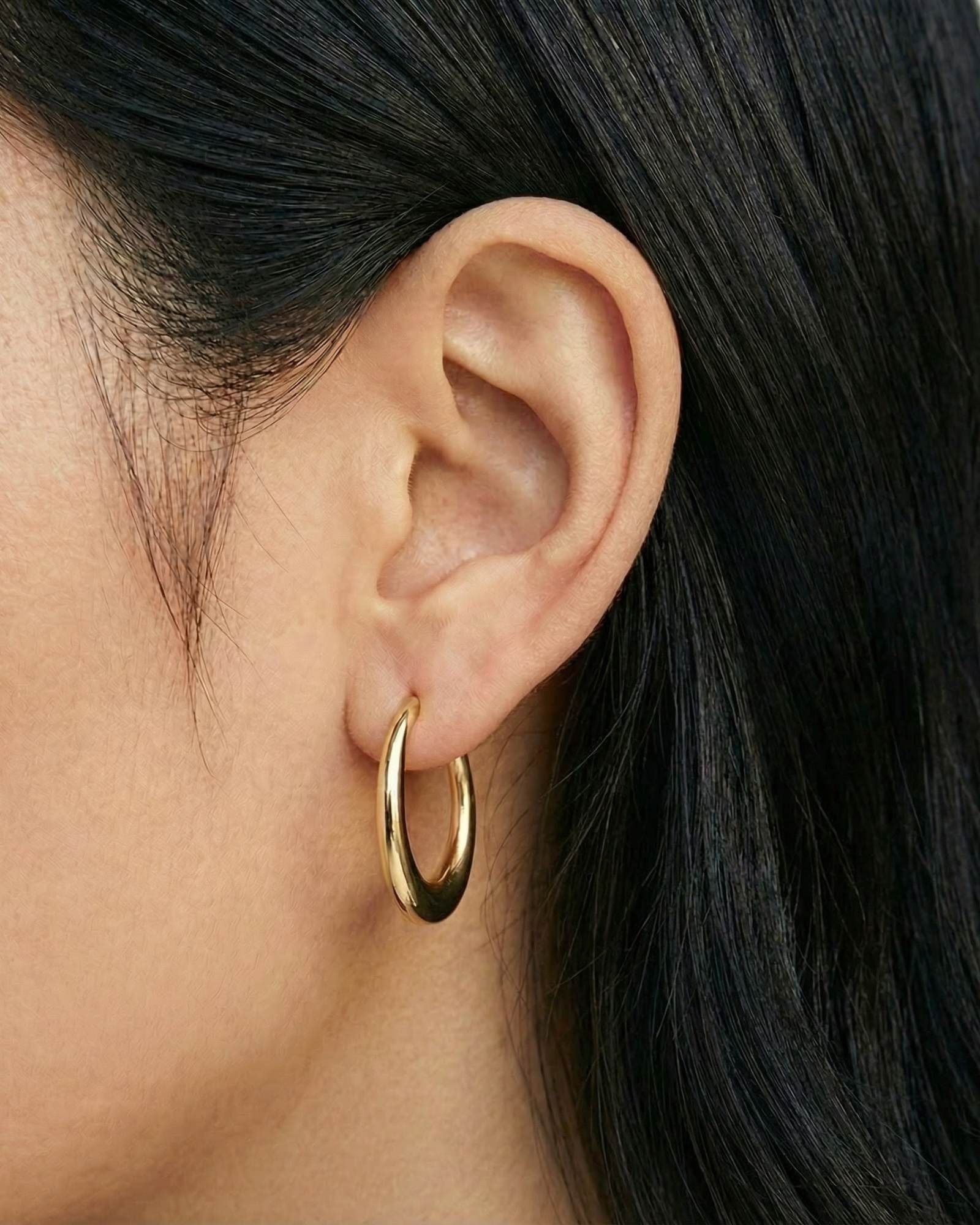 Close-up side profile of a woman’s ear wearing a single pair of polished gold hoop earrings with a rounded, medium-sized silhouette. The photo clearly shows the warm gold tone, clean finish, and proportion of the hoop on the ear against natural skin and dark hair. Designed for women seeking timeless, everyday gold hoop earrings, this image helps customers understand size, fit, and how classic gold hoops elevate a simple, minimal look.