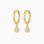 Pair of small gold huggie hoop earrings with suspended round bezel-set cubic zirconia charms, photographed on a clean neutral background. The polished 18k gold plated hoops have a smooth rounded shape and secure hinged closure, ideal for sensitive ears and everyday wear. Perfect for women looking for minimalist gold drop earrings that add refined sparkle without overpowering their look.