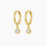 Pair of small gold huggie hoop earrings with suspended round bezel-set cubic zirconia charms, photographed on a clean neutral background. The polished 18k gold plated hoops have a smooth rounded shape and secure hinged closure, ideal for sensitive ears and everyday wear. Perfect for women looking for minimalist gold drop earrings that add refined sparkle without overpowering their look.