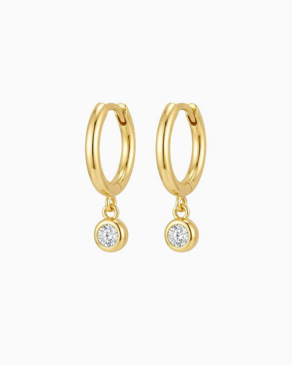 Pair of small gold huggie hoop earrings with suspended round bezel-set cubic zirconia charms, photographed on a clean neutral background. The polished 18k gold plated hoops have a smooth rounded shape and secure hinged closure, ideal for sensitive ears and everyday wear. Perfect for women looking for minimalist gold drop earrings that add refined sparkle without overpowering their look.