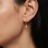 Close-up side profile of a woman wearing small gold huggie hoop earrings with a round bezel-set cubic zirconia drop charm. The lightweight 18k gold plated hoops sit snug on the lobe and feature a delicate dangling stone that adds subtle sparkle. Designed for women seeking minimal everyday jewellery, these dainty huggie earrings elevate a simple ear stack and offer an elegant solution for understated daily styling.