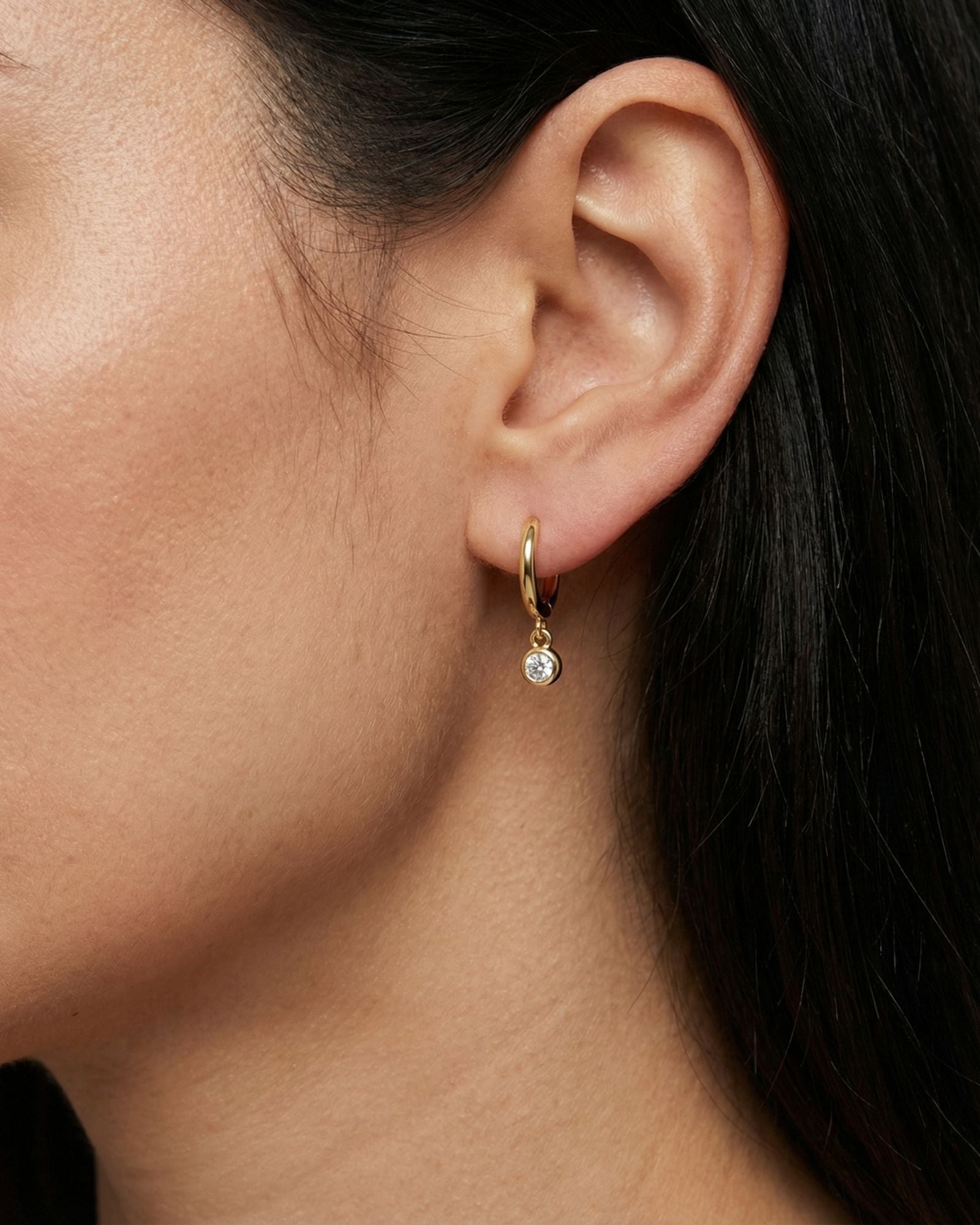 Close-up side profile of a woman wearing small gold huggie hoop earrings with a round bezel-set cubic zirconia drop charm. The lightweight 18k gold plated hoops sit snug on the lobe and feature a delicate dangling stone that adds subtle sparkle. Designed for women seeking minimal everyday jewellery, these dainty huggie earrings elevate a simple ear stack and offer an elegant solution for understated daily styling.