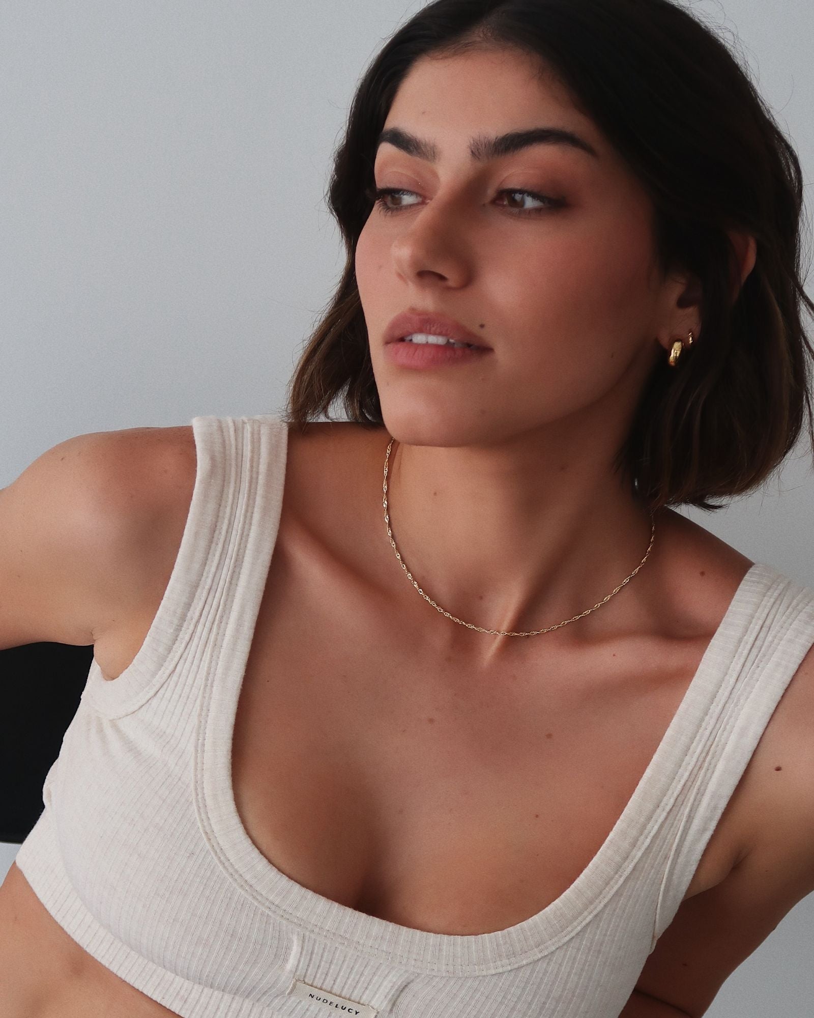 Lifestyle portrait of a woman wearing a delicate gold chain necklace styled close to the collarbone, paired with small gold hoop earrings and a neutral ribbed tank top. The image shows minimalist gold jewellery worn on bare skin in soft natural light, demonstrating how a fine gold necklace sits for everyday wear. Designed for women seeking understated, easy-to-style gold jewellery that complements casual outfits and solves the need for a lightweight, timeless necklace suitable for daily layering 