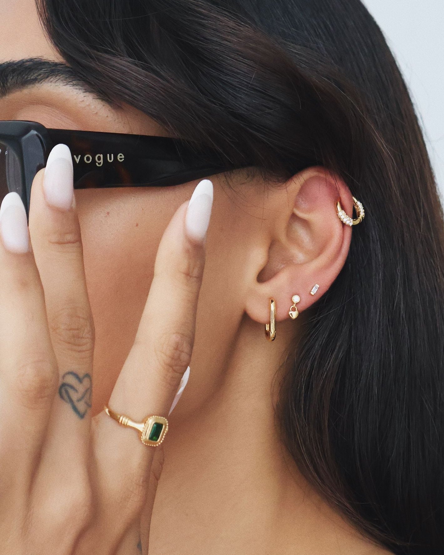 Close-up of a woman wearing black sunglasses and a layered gold ear stack, featuring small gold huggie hoop earrings in the lobe, a delicate gold charm stud, a slim gold helix hoop, and a pavé-style gold cartilage hoop. Styled with a gold ring set with a green rectangular stone, this image is for women looking for gold huggie earrings, cartilage and helix earrings, and modern ear stacking inspiration that is comfortable for everyday wear, suitable for sensitive ears, and ideal for creating a polished, fashi