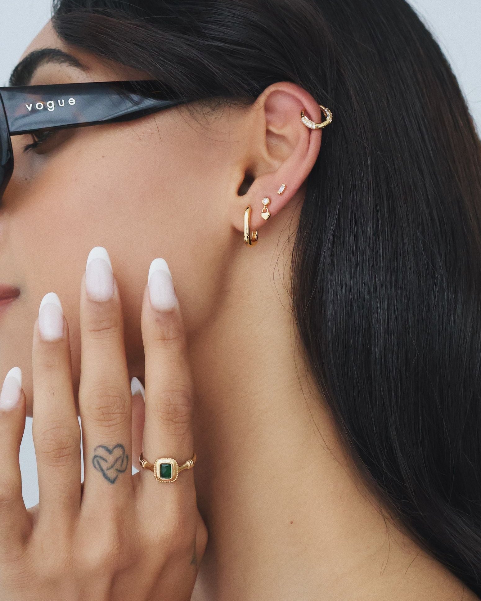 Side profile of a woman wearing a rectangular green emerald cubic zirconia gold ring on her finger, paired with layered gold hoop and stud earrings and a gold ear cuff. Styled for women seeking modern, vintage-inspired gold jewellery that adds colour and interest to everyday outfits while remaining minimal, wearable, and easy to layer.