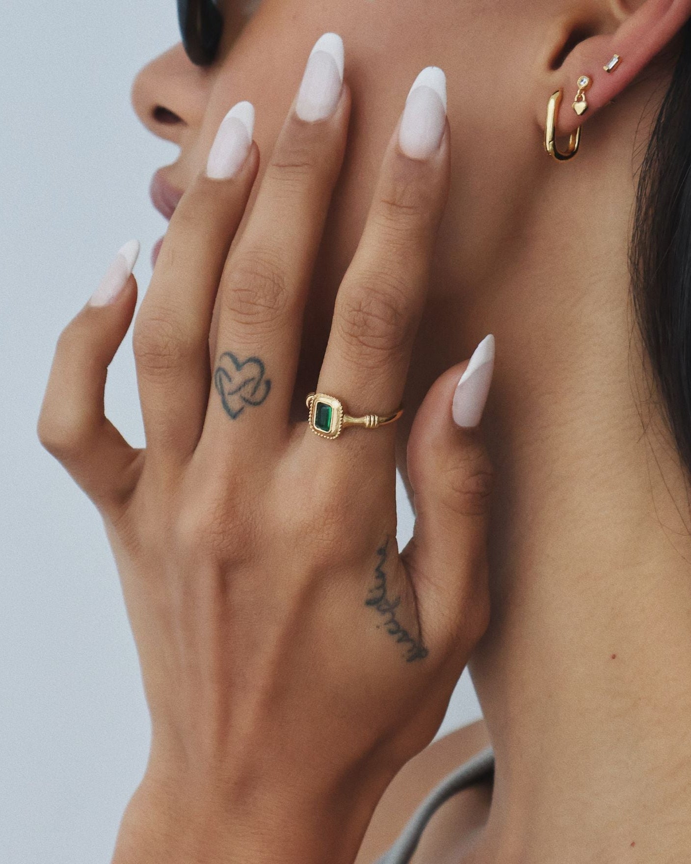 Close-up side profile of a woman wearing a rectangular green emerald cubic zirconia gold ring on her finger, paired with minimalist gold hoop and stud earrings. Designed for women looking for vintage-inspired gold rings that add colour and interest to everyday jewellery styling while remaining refined and wearable for daily use.