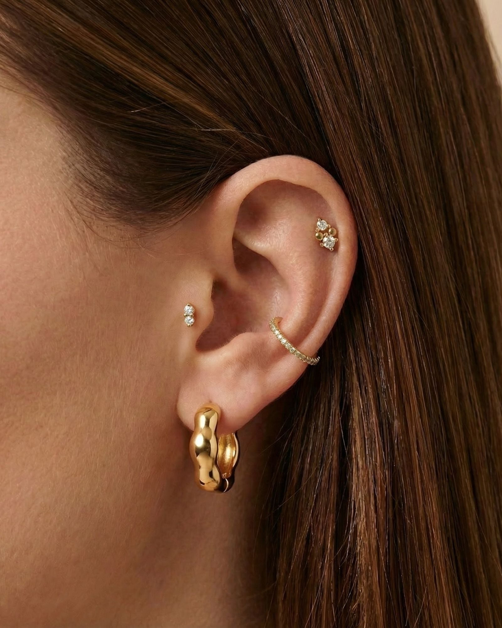 Close-up of a woman’s ear wearing Bella gold hoop earrings paired with a slim textured gold ear cuff and small crystal studs placed across the lobe and cartilage. This image is for women looking for gold hoop earrings that anchor a refined ear stack and show how to balance hoops, cuffs, and delicate studs for a cohesive everyday jewellery look.