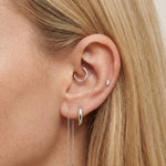 Close-up product photo of a woman’s ear with fair skin and straight blonde hair, styled with minimalist silver earrings. The ear features a small twisted silver hoop in the inner cartilage, a smooth silver huggie hoop in the lobe, a tiny marquise-shaped crystal stud in the upper ear, and a fine silver chain drop earring with two slim bar pendants. This image is for customers looking for understated silver earrings for everyday wear or multiple piercings. It demonstrates how delicate silver jewellery can be 
