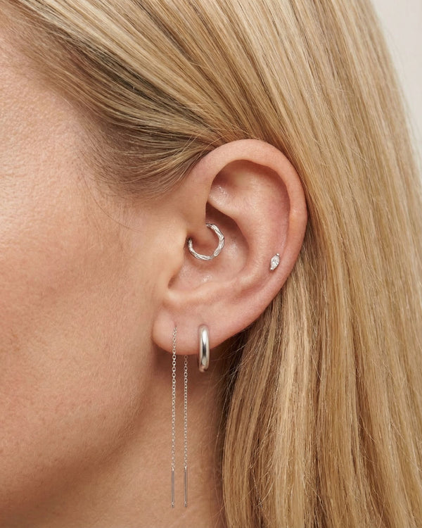 Close-up product photo of a woman’s ear with fair skin and straight blonde hair, styled with minimalist silver earrings. The ear features a small twisted silver hoop in the inner cartilage, a smooth silver huggie hoop in the lobe, a tiny marquise-shaped crystal stud in the upper ear, and a fine silver chain drop earring with two slim bar pendants. This image is for customers looking for understated silver earrings for everyday wear or multiple piercings. It demonstrates how delicate silver jewellery can be 