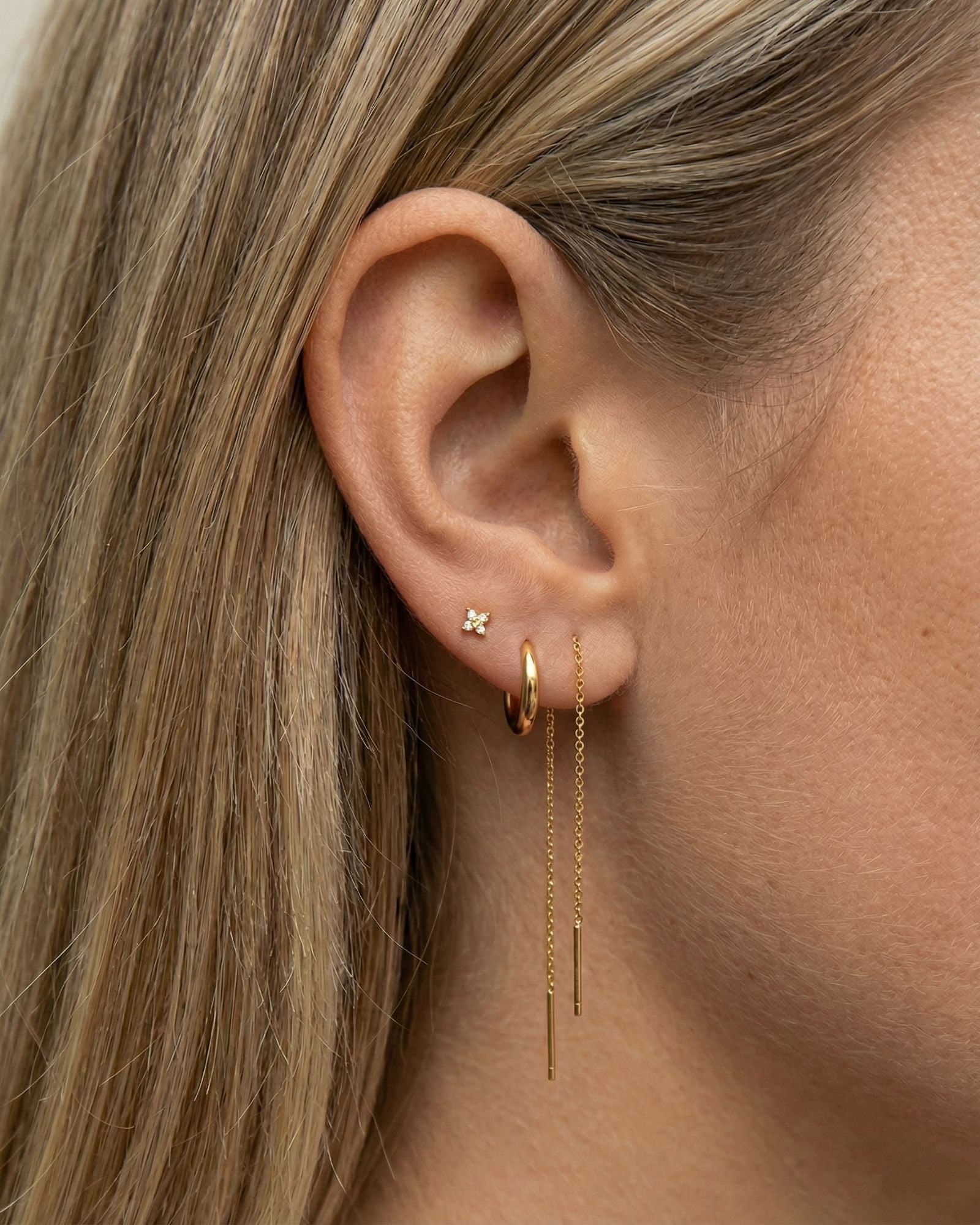 Close-up of a woman’s ear styled with a minimalist gold ear stack, featuring a dainty flower stud earring in the lobe, a small gold hoop, a delicate gold chain drop earring, and a tiny crystal flat-back stud in the upper cartilage. Designed for women seeking refined, everyday earrings for lobe and cartilage piercings, this image highlights comfortable, lightweight jewellery ideal for sensitive ears and effortless ear stacking.