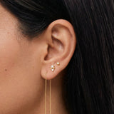 Close-up side profile of a woman’s ear wearing delicate gold earrings, featuring a fine double-chain threader earring with slim bar ends, paired with small gold stud earrings set with clear stones. Designed for women who prefer minimal, elegant gold jewellery, this image helps customers understand the length, movement, and layering of gold threader earrings when styled with classic stud piercings for everyday wear.
