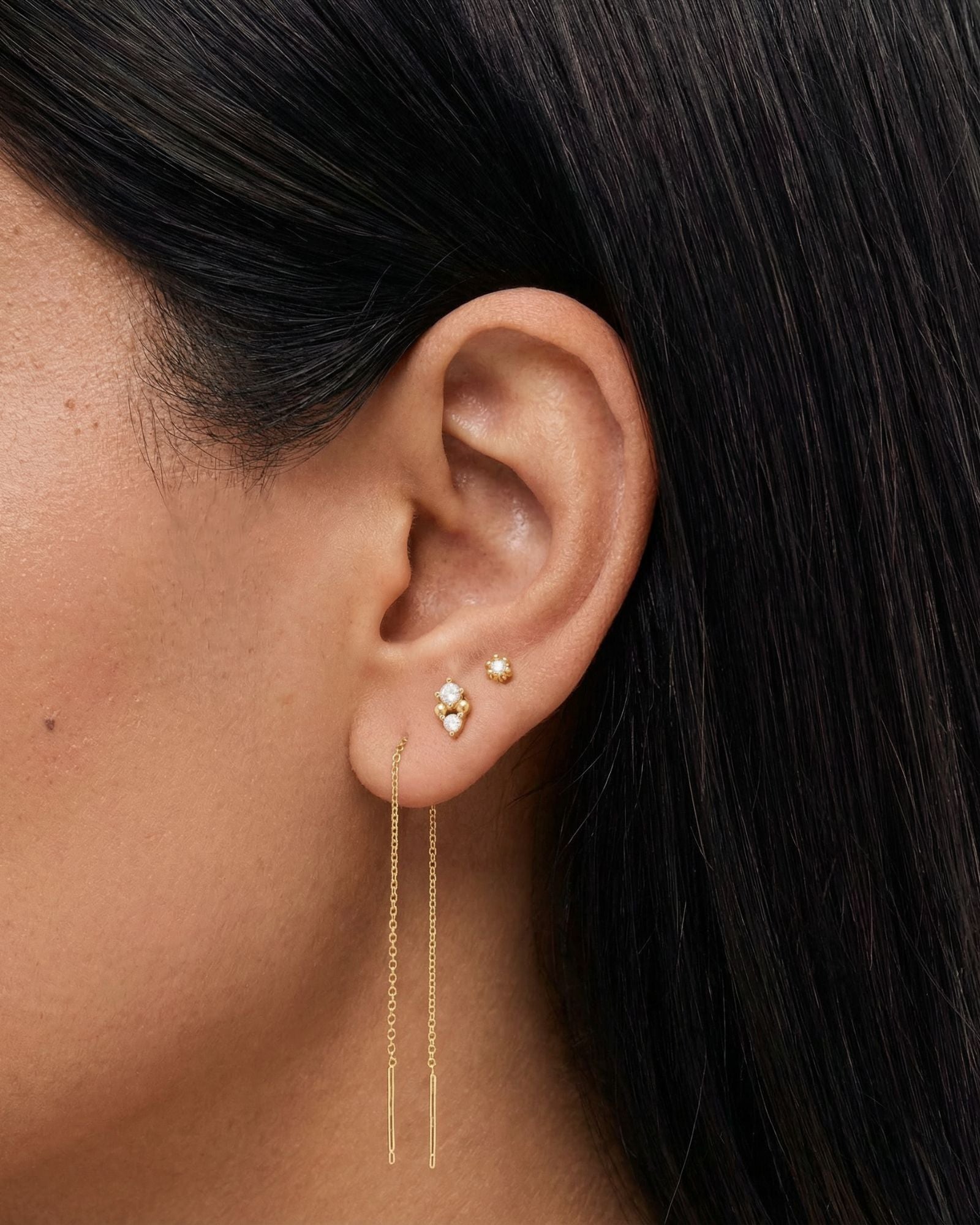 Close-up side profile of a woman’s ear wearing delicate gold earrings, featuring a fine double-chain threader earring with slim bar ends, paired with small gold stud earrings set with clear stones. Designed for women who prefer minimal, elegant gold jewellery, this image helps customers understand the length, movement, and layering of gold threader earrings when styled with classic stud piercings for everyday wear.