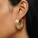 Close-up side profile of a woman wearing sculptural textured gold hoop earrings with a layered, organic ripple design in a high-polish gold finish. The statement hoops sit on the earlobe and feature a bold, modern silhouette. Designed for women who want elevated everyday jewellery or standout gold earrings for styling simple outfits, this image highlights how textured gold hoops add structure, dimension, and visual interest without stones. Ideal for those seeking contemporary statement earrings