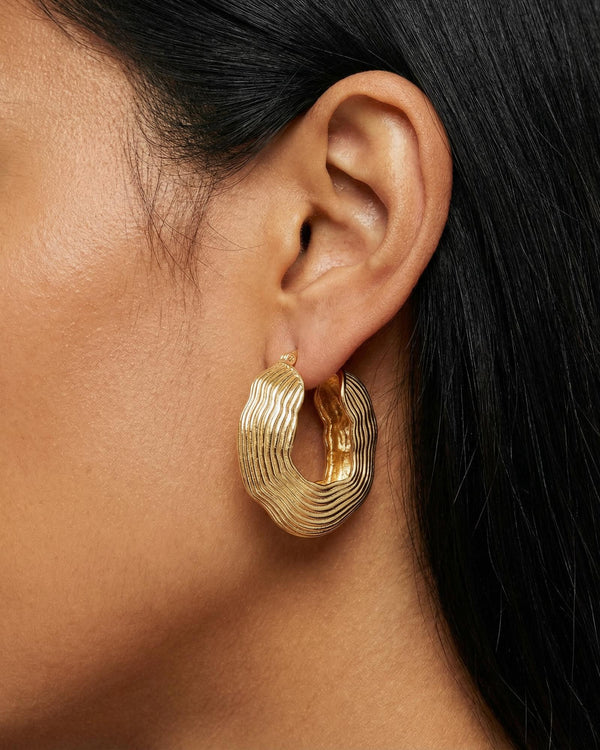 Close-up side profile of a woman wearing sculptural textured gold hoop earrings with a layered, organic ripple design in a high-polish gold finish. The statement hoops sit on the earlobe and feature a bold, modern silhouette. Designed for women who want elevated everyday jewellery or standout gold earrings for styling simple outfits, this image highlights how textured gold hoops add structure, dimension, and visual interest without stones. Ideal for those seeking contemporary statement earrings