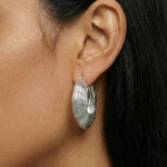 sculptural silver hoop earrings with a layered, organic ripple texture and high-polish finish. The statement earrings sit on the earlobe and feature a bold, fluid silhouette inspired by natural forms. Designed for women seeking modern silver statement earrings, this image shows how textured silver hoops add depth, structure, and visual interest to everyday outfits. Ideal for those looking for contemporary silver jewellery that elevates minimalist styling and provides an alternative to classic smooth hoops.