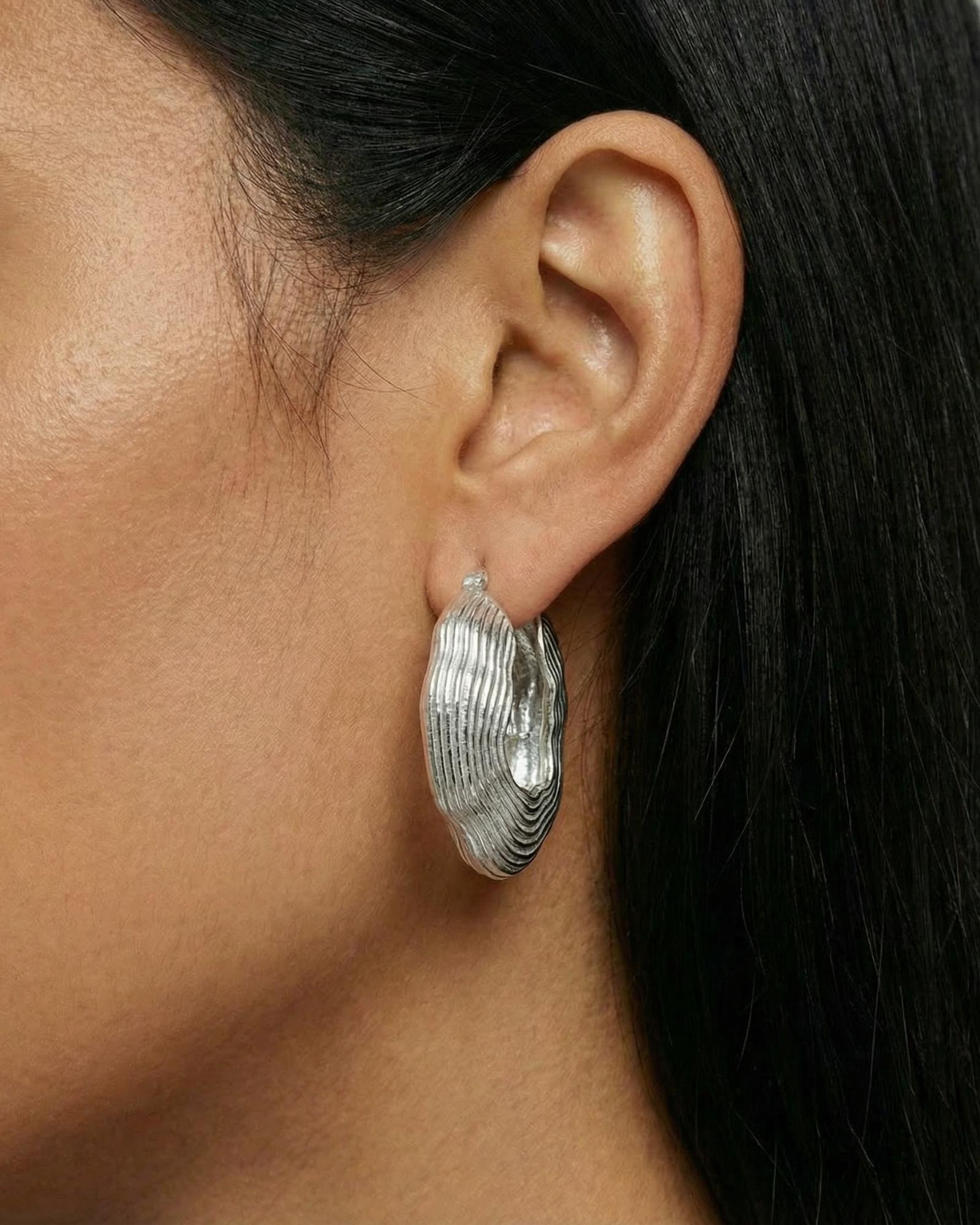 sculptural silver hoop earrings with a layered, organic ripple texture and high-polish finish. The statement earrings sit on the earlobe and feature a bold, fluid silhouette inspired by natural forms. Designed for women seeking modern silver statement earrings, this image shows how textured silver hoops add depth, structure, and visual interest to everyday outfits. Ideal for those looking for contemporary silver jewellery that elevates minimalist styling and provides an alternative to classic smooth hoops.