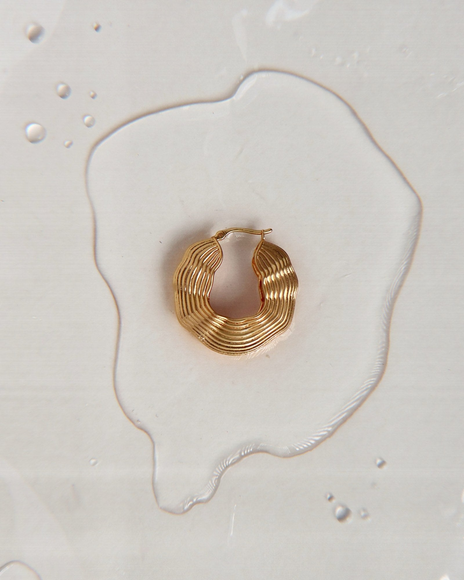 Gold sculptural statement hoop earring photographed on a wet surface with water droplets, highlighting its textured layered design in an editorial product shot.
