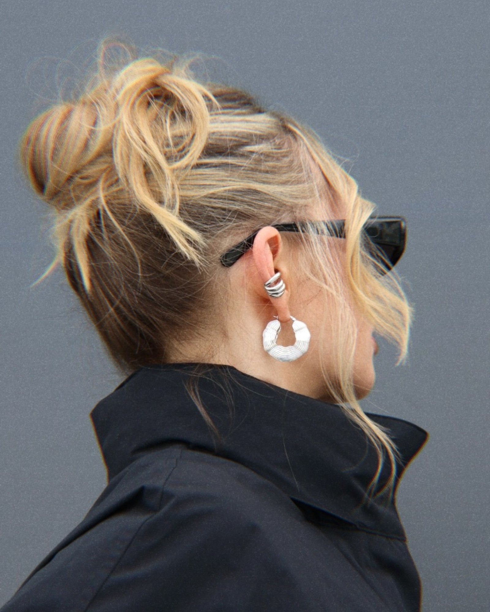 Side profile of a woman wearing large silver sculptural statement hoop earrings styled with a silver ear cuff, black sunglasses and a black coat for a modern street-style look