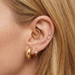 Close-up of a woman’s ear styled with multiple gold earrings. The ear features a thick, smooth gold huggie in the lobe, a smaller gold hoop with a tiny star-shaped diamond charm beside it, a textured gold hoop in the upper cartilage, and two small gold studs with delicate diamonds placed vertically along the helix. The model’s hair is tucked behind the ear, highlighting the curated gold stack.